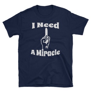 May include: Navy blue t-shirt with the white text "I Need A Miracle" and a graphic of a hand with the index finger pointing up. The shirt is made of a soft material and has short sleeves.