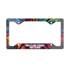 May include: License plate frame with a colorful, abstract design featuring shades of blue, orange, and purple. The frame has the text "WHAT A LONG STRANGE TRIP ITS BEEN" in white.