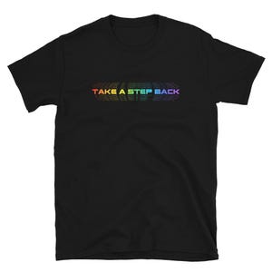 Puede incluir: Camiseta negra con el texto "TAKE A STEP BACK" en un degradado arcoíris en el pecho. El texto se repite varias veces, creando un efecto de capas. La camiseta tiene mangas cortas y cuello redondo.