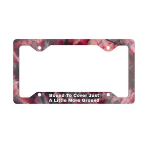 May include: Rectangular license plate frame with a colorful, abstract design in shades of red, pink, and gray. The frame has four mounting holes and the text "Bound To Cover Just A Little More Ground" in white.