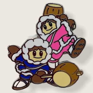 May include: A pin featuring two cartoon characters wearing winter clothing. The character on top is wearing a pink jacket and a brown hat. The character on the bottom is wearing a blue jacket and a white hat. Both characters are holding a brown stick.