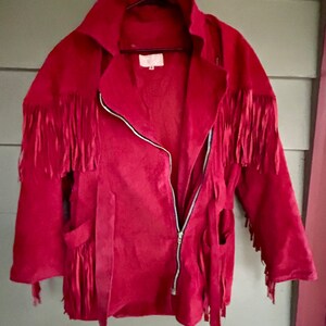 Puede incluir: Una chaqueta de ante roja con detalles de flecos en los hombros, las mangas y los bolsillos. La chaqueta tiene una cremallera y un cinturón.