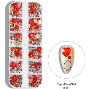 Op de afbeelding: Een doorzichtige plastic container met 10 vakken gevuld met rode en zilveren nagelkunstversieringen. De versieringen omvatten harten, ringen en strasssteentjes. De container is gelabeld "CapreDiemNail B-06".