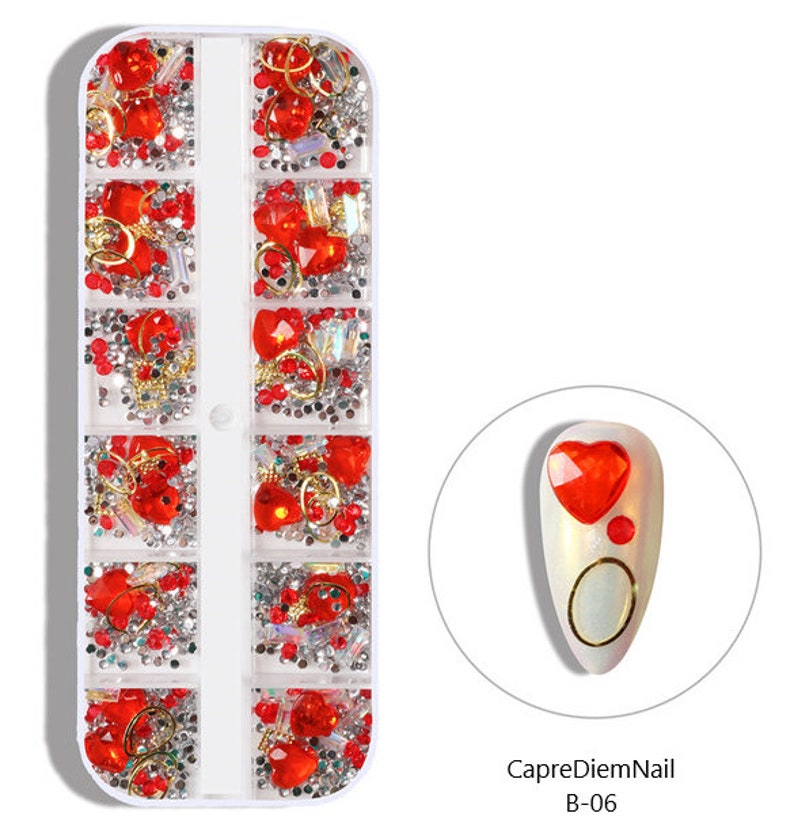 Op de afbeelding: Een doorzichtige plastic container met 10 vakken gevuld met rode en zilveren nagelkunstversieringen. De versieringen omvatten harten, ringen en strasssteentjes. De container is gelabeld "CapreDiemNail B-06".