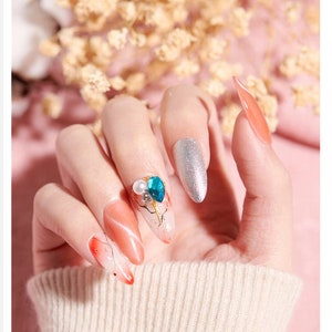 Op de afbeelding: Een handmodel met lange, amandelvormige nagels. De nagels zijn geverfd met een gradi&euml;nt van oranje en wit, en &eacute;&eacute;n nagel heeft een blauw en wit strass-ontwerp.
