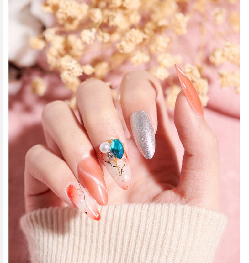 Op de afbeelding: Een handmodel met lange, amandelvormige nagels. De nagels zijn geverfd met een gradi&euml;nt van oranje en wit, en &eacute;&eacute;n nagel heeft een blauw en wit strass-ontwerp.