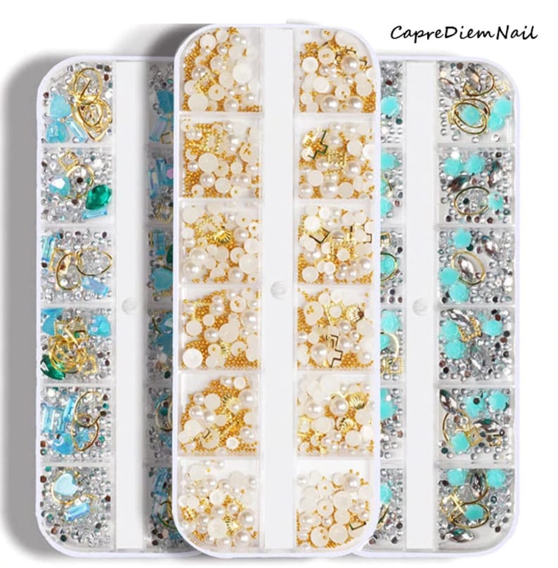 Op de afbeelding: Drie witte plastic containers gevuld met verschillende nagelkunstversieringen. De containers zijn elk in 12 secties verdeeld. De versieringen omvatten parels, strasssteentjes en kleine gouden en zilveren bedels.