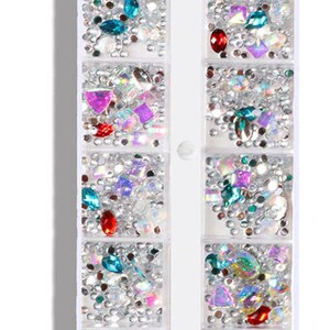 Op de afbeelding: Een doorzichtige plastic container met 12 vakken gevuld met verschillende gekleurde strass steentjes en glitter. De container is gelabeld "CapreDiemNail B-03".