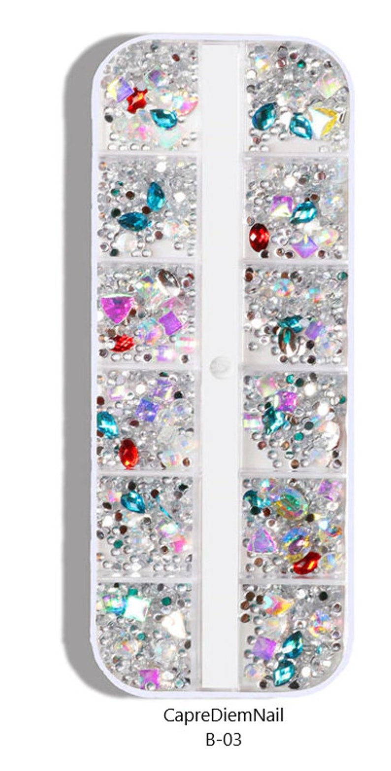 Op de afbeelding: Een doorzichtige plastic container met 12 vakken gevuld met verschillende gekleurde strass steentjes en glitter. De container is gelabeld "CapreDiemNail B-03".