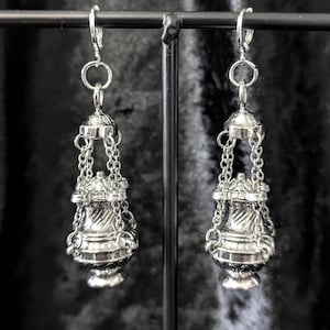 Peut inclure: Une paire de boucles d'oreilles pendantes en argent avec un design détaillé et orné. Chaque boucle d'oreille présente un petit pendentif rond avec une chaîne et un pendentif plus grand en forme de cloche avec une chaîne.
