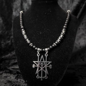 Puede incluir: Un collar que presenta una serie de cuentas esféricas de color negro mate intercaladas con pequeños espaciadores de metal plateado. La pieza central es un gran colgante de metal plateado con un complejo diseño geométrico de sigilo.