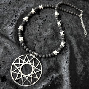 Könnte beinhalten: Eine Halskette mit einem großen, kreisförmigen, silberfarbenen Anhänger mit einem komplexen geometrischen Sternmuster. Die Kette besteht aus dunklen, texturierten Lavaperlen, die mit kleinen, weißen, knochenartigen Elementen und einer silberfarbenen Verlängerungskette versehen sind.