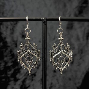 May include: A pair of silver-colored filigree earrings. Each earring features an ornate, symmetrical design with intricate cut-out patterns and a dangling hook. The earrings are displayed against a dark, textured background.
