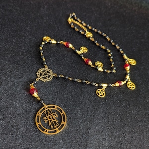 Könnte beinhalten: Eine goldfarbene Halskette mit kleinen schwarzen Perlen, roten Akzentperlen und mehreren goldenen Pentagramm-Anhängern. Die Kette hat einen zentralen runden Anhänger mit einem komplexen Siegelmotiv und ein kleineres Pentagramm darüber.