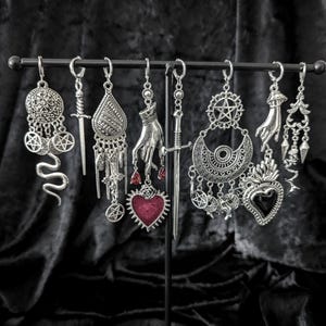Op de afbeelding: Een verzameling zilverkleurige oorbellen met gotische en occulte thema's. Ontwerpen omvatten dolken, handen, harten, pentagrammen en slangenmotieven. De oorbellen worden tentoongesteld op een zwarte metalen standaard.