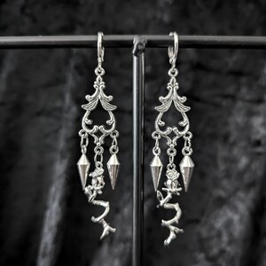 Op de afbeelding: Zilverkleurige oorhangers met sierlijke filigraan toppen, hangende geometrische vormen en takachtige accenten. Deze oorbellen hebben een gotische of Victoriaanse stijl, perfect om een vleugje elegantie en edge aan elke outfit toe te voegen.