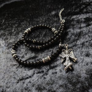 Op de afbeelding: Een kralenketting met kleine, ronde, gepolijste zwarte kralen afgewisseld met grotere, grijze, getextureerde bolvormige kralen en zilverkleurige metalen tussenstukjes. Een zilverkleurige hanger in de vorm van een gevleugelde, gehoornde figuur hangt aan de ketting.