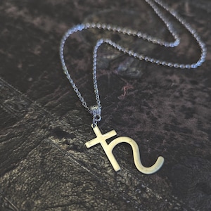 Op de afbeelding: Een zilverkleurige metalen ketting met een hanger in de vorm van het astrologische symbool voor Saturnus. De hanger hangt aan een fijne zilverkleurige ketting met kleine ronde schakels en heeft een gladde, gepolijste afwerking op het metaal.
