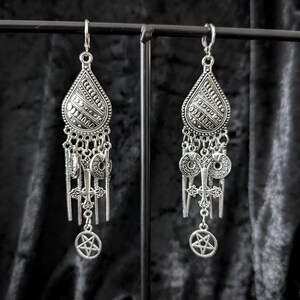 Op de afbeelding: Een paar zilverkleurige oorhangers. Elke oorbel heeft een druppelvormige bovenkant met ingewikkelde patronen, verbonden met meerdere bedels, waaronder kruisen en pentagrammen. De oorbellen worden tegen een donkere achtergrond gepresenteerd.