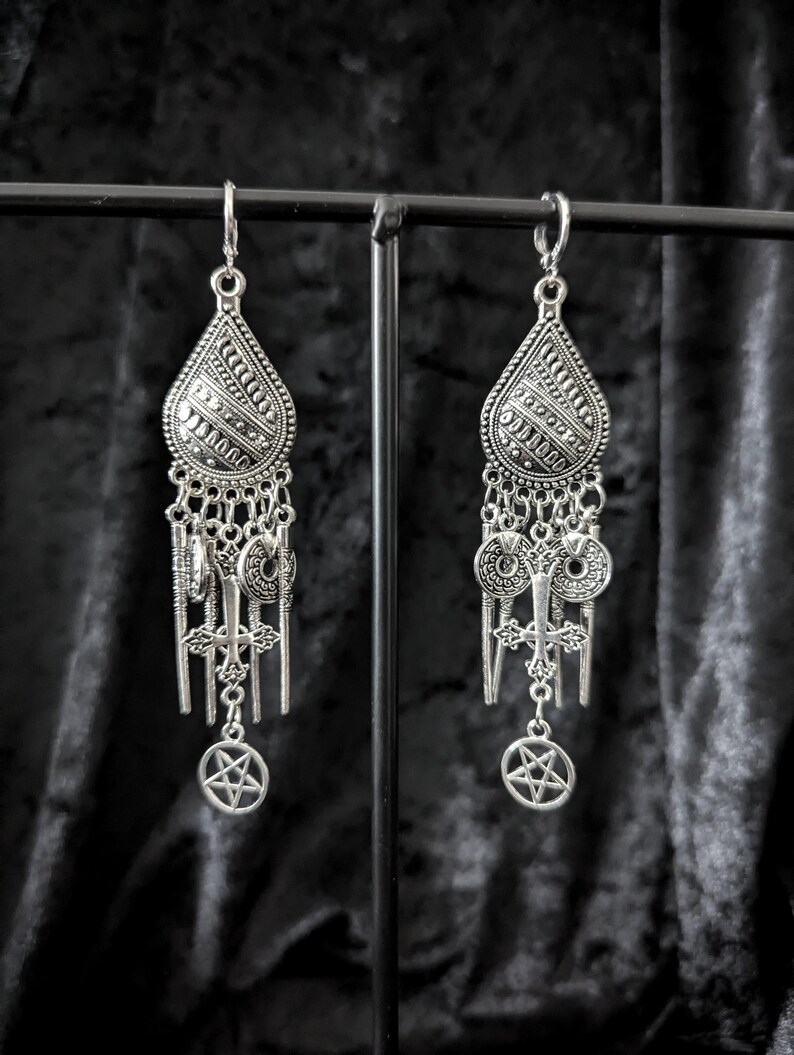 Op de afbeelding: Een paar zilverkleurige oorhangers. Elke oorbel heeft een druppelvormige bovenkant met ingewikkelde patronen, verbonden met meerdere bedels, waaronder kruisen en pentagrammen. De oorbellen worden tegen een donkere achtergrond gepresenteerd.