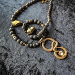Puede incluir: Un conjunto de collar y pulsera con cuentas de lava porosas de color gris oscuro, detalles de cabezas de serpiente de metal en tono dorado antiguo y espaciadores decorativos. El collar incluye un colgante de metal dorado con forma de serpiente.