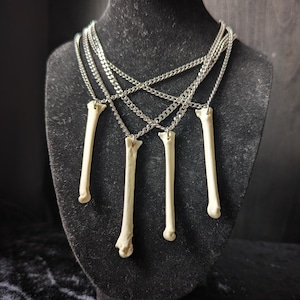 Puede incluir: Un collar de cadenas múltiples en tono plateado con cuatro colgantes de hueso alargados en color blanco roto que cuelgan a diferentes alturas. Las cadenas metálicas están interconectadas, creando un efecto drapeado con los huesos.