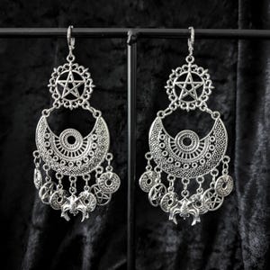 Op de afbeelding: Zilverkleurige oorhangers met een gotisch thema. Elke oorbel heeft een pentagram hanger, een halve maan vorm en bungelende bedels met vleermuis- en cirkelontwerpen. De oorbellen worden gepresenteerd tegen een donkere achtergrond.