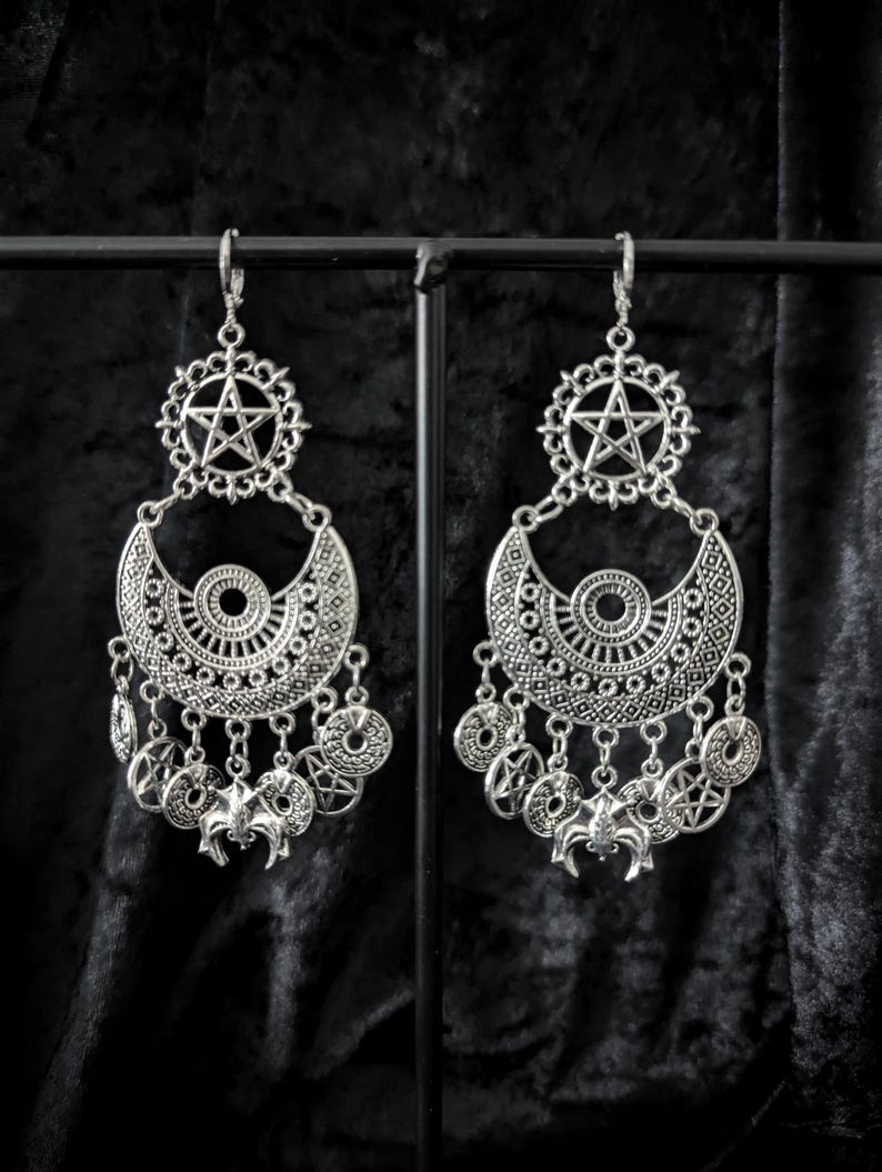 Op de afbeelding: Zilverkleurige oorhangers met een gotisch thema. Elke oorbel heeft een pentagram hanger, een halve maan vorm en bungelende bedels met vleermuis- en cirkelontwerpen. De oorbellen worden gepresenteerd tegen een donkere achtergrond.