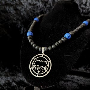 May include: A necklace featuring a silver pendant with a detailed design, suspended from a strand of black lava beads interspersed with blue beads. The necklace is displayed on a black velvet stand, set against a dark background.