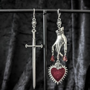 Op de afbeelding: Zilverkleurige oorhangers. E&eacute;n oorbel heeft een zwaard, de andere een hand met rode druppelaccenten en een hartvormige hanger met een oogontwerp. Het hart is rood met een zilveren rand. De oorbellen hangen aan een zwarte metalen display.