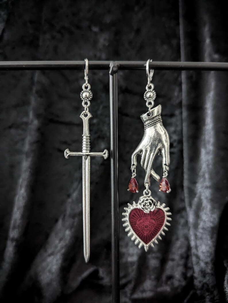 Op de afbeelding: Zilverkleurige oorhangers. E&eacute;n oorbel heeft een zwaard, de andere een hand met rode druppelaccenten en een hartvormige hanger met een oogontwerp. Het hart is rood met een zilveren rand. De oorbellen hangen aan een zwarte metalen display.