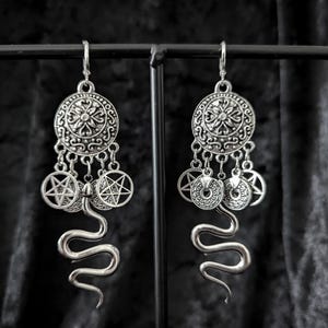 Op de afbeelding: Zilverkleurige oorhangers met een gotisch thema. Elke oorbel heeft een cirkelvormige hanger met ingewikkelde details, bungelende bedels met pentagrammen en een gestileerde slang. De oorbellen worden tegen een donkere achtergrond gepresenteerd.
