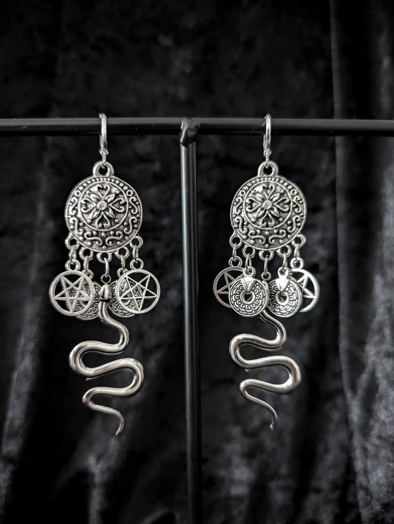 Op de afbeelding: Zilverkleurige oorhangers met een gotisch thema. Elke oorbel heeft een cirkelvormige hanger met ingewikkelde details, bungelende bedels met pentagrammen en een gestileerde slang. De oorbellen worden tegen een donkere achtergrond gepresenteerd.