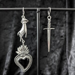 Op de afbeelding: Zilverkleurige oorbellen met een gotisch thema. E&eacute;n oorbel heeft een hand die een vlammend hart met een zwart midden vasthoudt. De andere oorbel is een dolk met een schedel op het handvat. De oorbellen hangen aan een zwart metalen display.