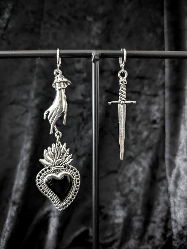 Op de afbeelding: Zilverkleurige oorbellen met een gotisch thema. E&eacute;n oorbel heeft een hand die een vlammend hart met een zwart midden vasthoudt. De andere oorbel is een dolk met een schedel op het handvat. De oorbellen hangen aan een zwart metalen display.