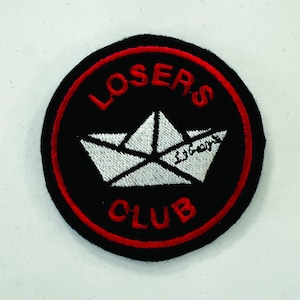 May include: A round, black patch with a red border. The patch features the words "LOSERS CLUB" in red, arched around the top and bottom. A white paper boat is in the center, with text in a foreign language. This patch is likely for clothing or accessories.