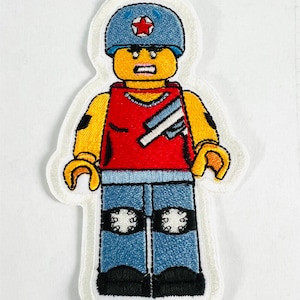 May include: Embroidered patch of a Lego minifigure wearing a blue helmet with a red star, a red shirt with white stripes, blue jeans with white knee pads, and black shoes.