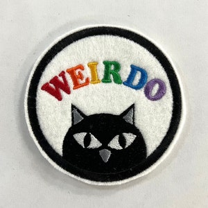 May include: A round, white, embroidered patch with a black outline. The word "WEIRDO" is written in rainbow colors above a black cat with white eyes.