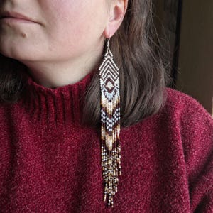 May include: Long beaded earrings with a geometric design. The earrings feature a pattern of white, brown, and gold beads. The earrings are hanging from a silver hook and are worn by a person wearing a red sweater.