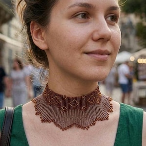 Op de afbeelding: Een kralen choker-halsketting met een dichte, donker koperkleurige geweven band met een herhalend ruitpatroon. De onderrand van de ketting loopt uit in een delicate, geschulpte franje van fijne, bungelende kralen in een bijpassende tint.