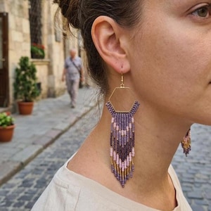 May include: Gold hexagon earrings with beaded fringe in purple, pink, and gold. These statement earrings feature a geometric design and a bohemian style. The beaded fringe adds texture and colour. The earrings are suitable for various occasions.
