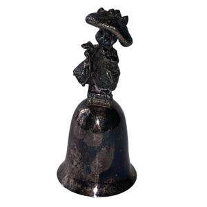 May include: A silver-toned bell with a figure of a person wearing a hat and carrying a bag. The bell has a flared base and a detailed figure on top, likely a collectible or decorative item.