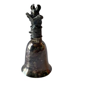May include: Silver-toned bell with a detailed figure of a rabbit reading a book on top. The bell has a flared base and a textured surface, reflecting light. A unique decorative item.