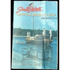 May include: A 2003 South Dakota Official Highway Map with a boat illustration on the cover. The map features the text "South Dakota" in red script, and the phrase "Great Faces, Great Places" at the bottom.