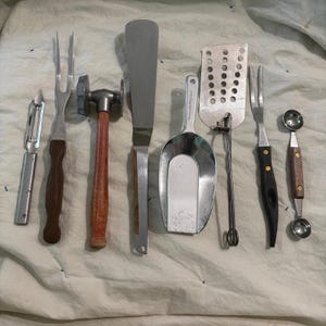 May include: A collection of kitchen utensils including a vegetable peeler, a two-pronged fork with a brown handle, a meat tenderizer with a wooden handle, a long metal spatula, a metal scoop, a slotted turner, a fork with a black handle, and a melon baller.