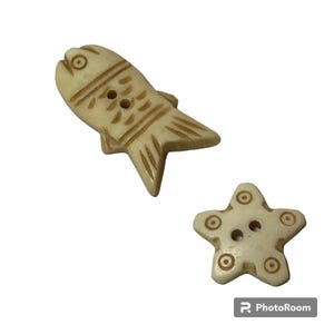 Puede incluir: Dos botones decorativos: uno con forma de pez y otro con forma de estrella de mar. Ambos botones son de color blanquecino con detalles tallados. El botón de pez tiene dos agujeros para coser y líneas grabadas. La estrella de mar tiene cinco puntas con detalles circulares y dos agujeros.