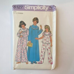 May include: Simplicity pattern 8127 for girls and chubbies. The pattern features illustrations of three girls in pajamas. One girl is holding a cat, another is wearing a blue nightgown, and the third is wearing a floral pajama set.