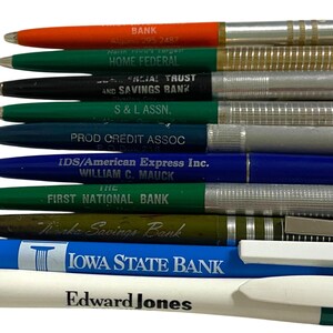 May include: A collection of ballpoint pens in various colors, including orange, green, black, and blue. Each pen is imprinted with a different bank or financial institution's name, such as "HOME FEDERAL" and "IOWA STATE BANK". The pens have silver-colored metal accents.
