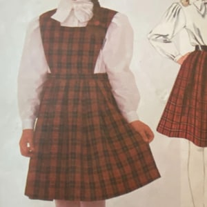Può includere: Un abito a trapezio a quadri rossi, marroni e neri, abbinato a una camicetta bianca. L'abito presenta uno scollo quadrato e una gonna a pieghe. Lo stile suggerisce una classica uniforme scolastica o moda vintage.