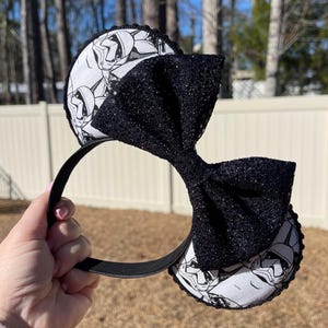 May include: Black headband with two round ears featuring a black and white Stormtrooper design. A large, sparkly black bow is centered between the ears. The headband is held by a hand against a blurred outdoor background.